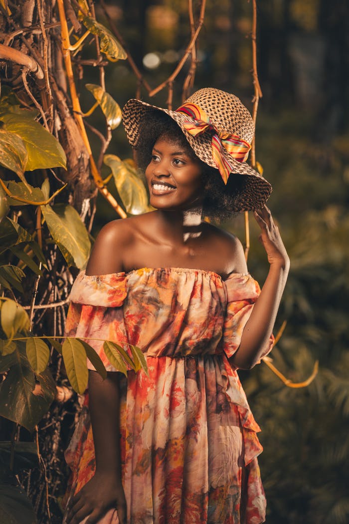 A beautiful woman in a floral dress and hat poses gracefully outdoors.